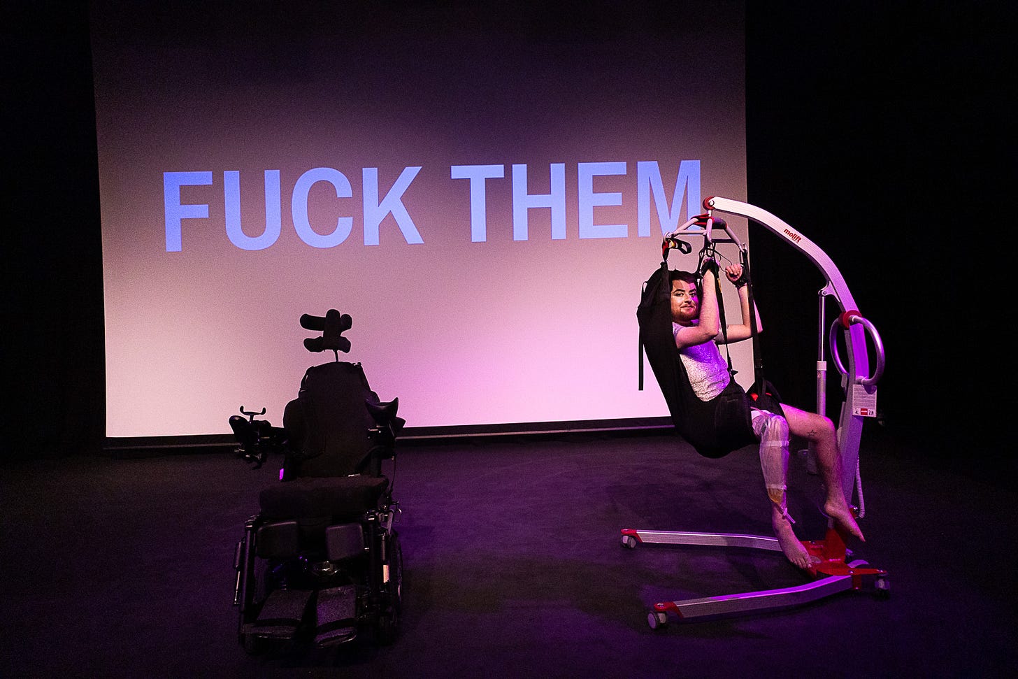 Image: Jamie Hale, a white person with dark red hair and a ginger beard is in a hoist sling, wearing a silver shirt and stockings, catheter bag on their legs. Their hands are handcuffed to the hoist. Beside them is their electric wheelchair, empty. Behind them is the text “FUCK THEM”. Jamie has a cheeky look on their face.