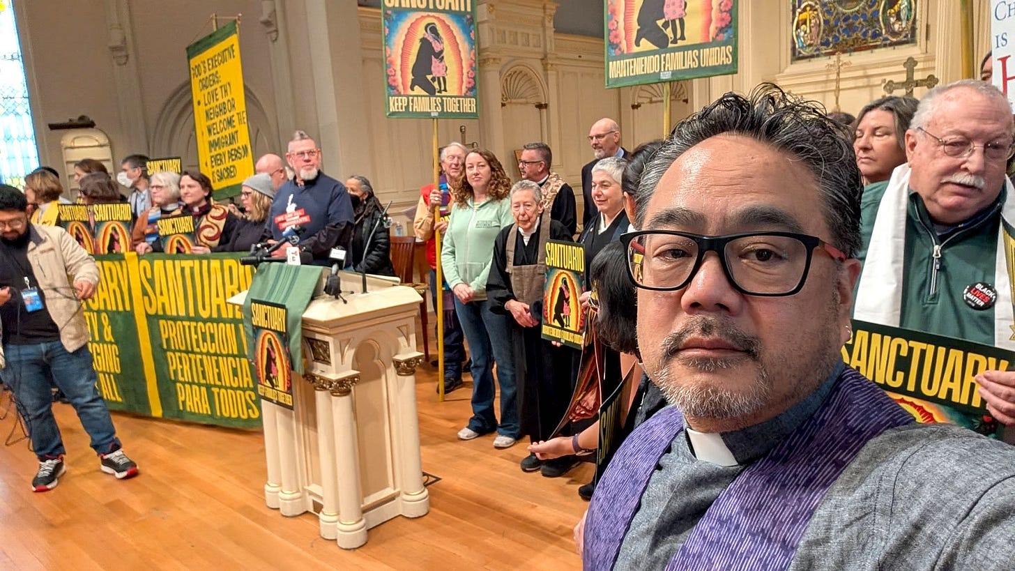 Bruce is in the foregraound of a large gathering of multifath leaders inside a church sanctuary. They carry signs advocating SANCTUARY and immigration justice.