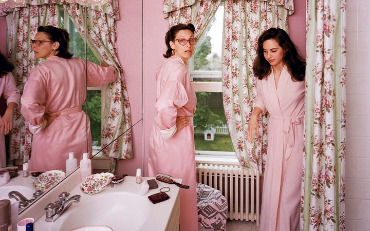 1: Photograph by Tina Barney depicting a bed covered in a white embroidered bedspread adorned with pink flowers. A young girl in a white dress with puffed sleeves and her hair adorned with ribbons sits on the bed. Next to her is a man in a blue shirt, leaning on the bed, appearing to engage in conversation with her. In the background, another girl in a pastel-colored dress stands in a brightly lit closet filled with clothes hanging on a rod. 2: Photograph by Tina Barney depicting two women in a room with cream colored walls. On the left, one woman with wavy brown hair is seated, turned toward the camera, resting her chin on her hand. To the right, facing a mirror, a young woman with short, bleach blonde hair applies bright red lipstick. She is also dressed in black and stands in a profile position, reflecting in the mirror. 3: Photograph by Tina Barney, depicting three people engaged in conversation in a sunlit room. On the left, an older man with white hair wearing a black vest over a white shirt gestures towards the center. In the middle, a young person stands with their hands holding the back of their neck. To the right, a woman in a light brown blouse and white skirt holds her chin thoughtfully. 4: Photograph by Tina Barney featuring two women in a pastel pink bathroom, both wearing pink robes. On the left, one woman with short dark hair and glasses gazes toward the camera, her hand on her hip. She is standing in front of a mirror, in which her reflection is visible. To the right, a young woman with long dark hair peeks from behind floral curtains covering a window.