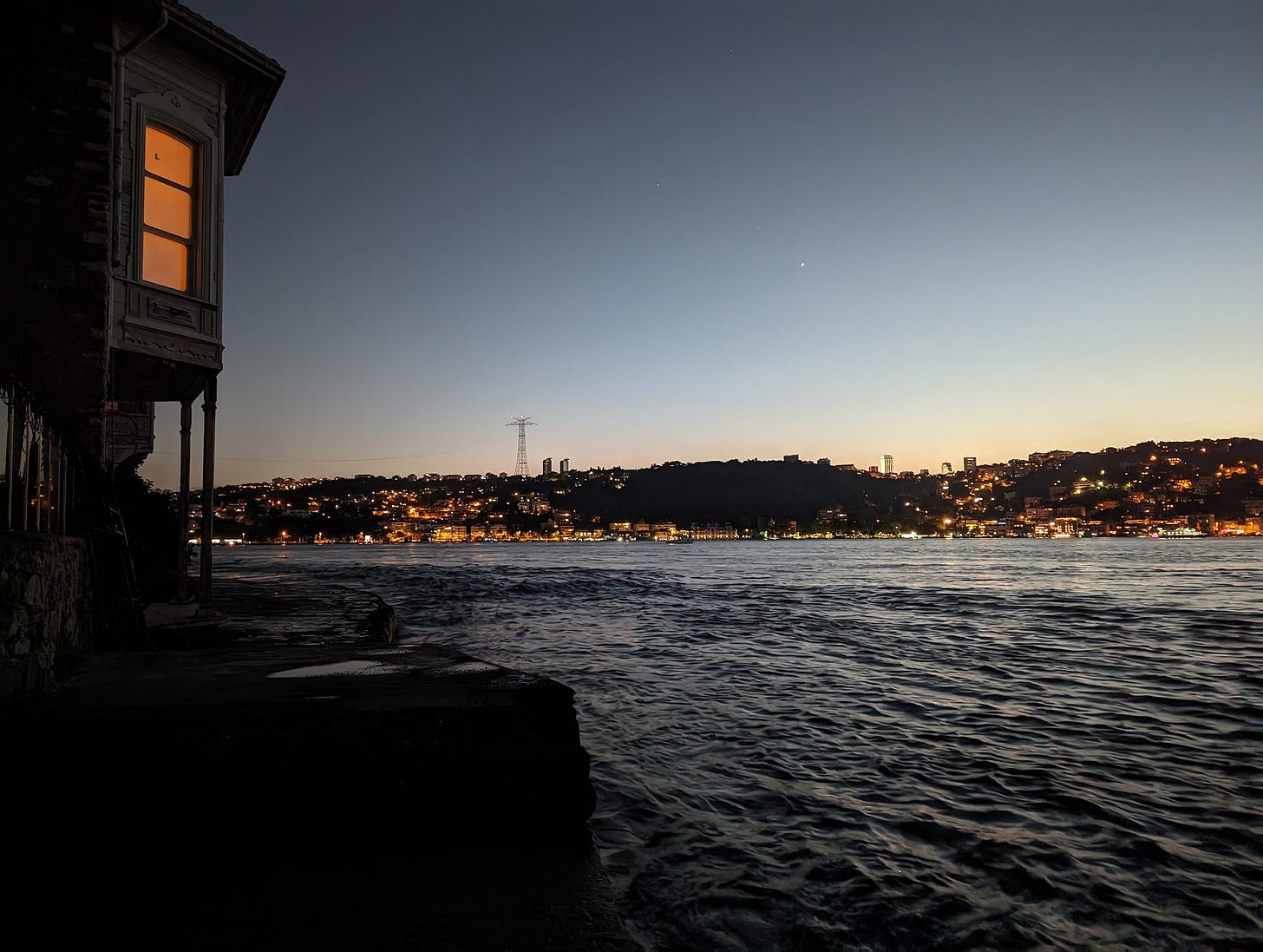 Bir Boğaz fotosu, akşam. Solda karanlıkta bir yalınin penceresinde sarı ışık. Fotoğrafın kalanında Boğaz ve karşı yaka görünüyor. Akıntı var. Su karanlık. 