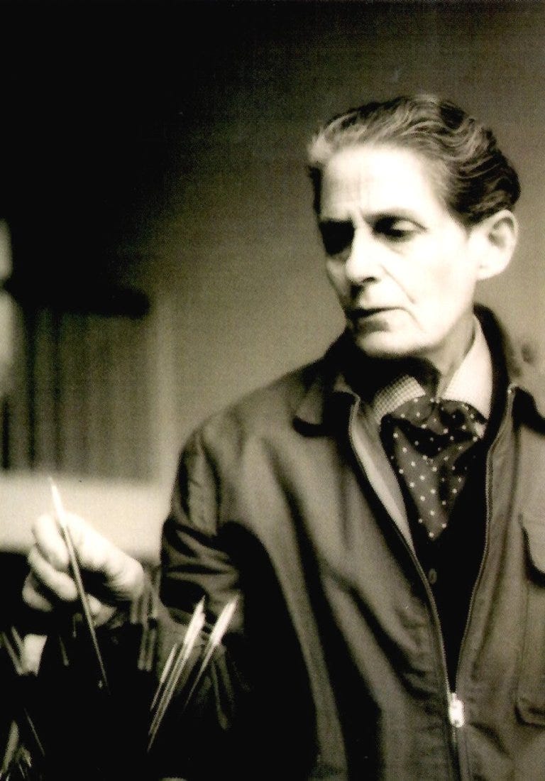 Black and white picture of Gluck as an elderly person with slicked-back silver and black hair, wearing a dark jacket and polka dot scarf, focuses intently on paintbrushes in a cup in front of them in a softly lit setting. Gluck has a serious, contemplative look on their face. Black and white picture of Gluck as an elderly person with slicked-back silver and black hair, wearing a dark jacket and polka dot scarf, focuses intently on paintbrushes in a cup in front of them in a softly lit setting. Gluck has a serious, contemplative look on their face.