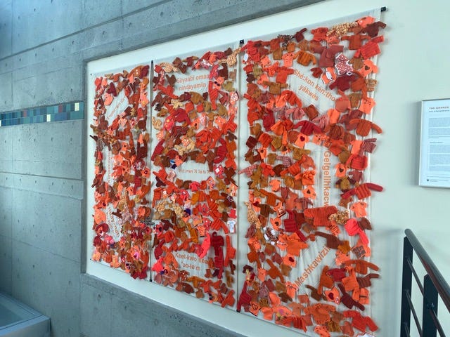 A three‑panel installation mounted on a light-colored wall, each panel densely covered with hundreds of small handcrafted orange sweaters and clothing pieces in shades ranging from bright orange to deep rust. The items are knitted, crocheted, or sewn, creating a textured and layered surface across all three panels. Beneath the sweaters, white fabric panels display faint printed text partially visible through gaps in the textile pieces. The installation is displayed in a concrete stairwell with natural light filtering from the left, and a small wall placard with project information is mounted to the right of the artwork. A three‑panel installation mounted on a light-colored wall, each panel densely covered with hundreds of small handcrafted orange sweaters and clothing pieces in shades ranging from bright orange to deep rust. The items are knitted, crocheted, or sewn, creating a textured and layered surface across all three panels. Beneath the sweaters, white fabric panels display faint printed text partially visible through gaps in the textile pieces. The installation is displayed in a concrete stairwell with natural light filtering from the left, and a small wall placard with project information is mounted to the right of the artwork.