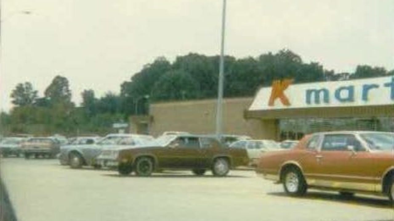 A vintage photo of a crowded Kmart parking lot, likely from the 1970s or 1980s, with classic cars parked in front of the store’s iconic red and blue signage. The image captures a nostalgic moment of American retail culture and consumer life at its peak.