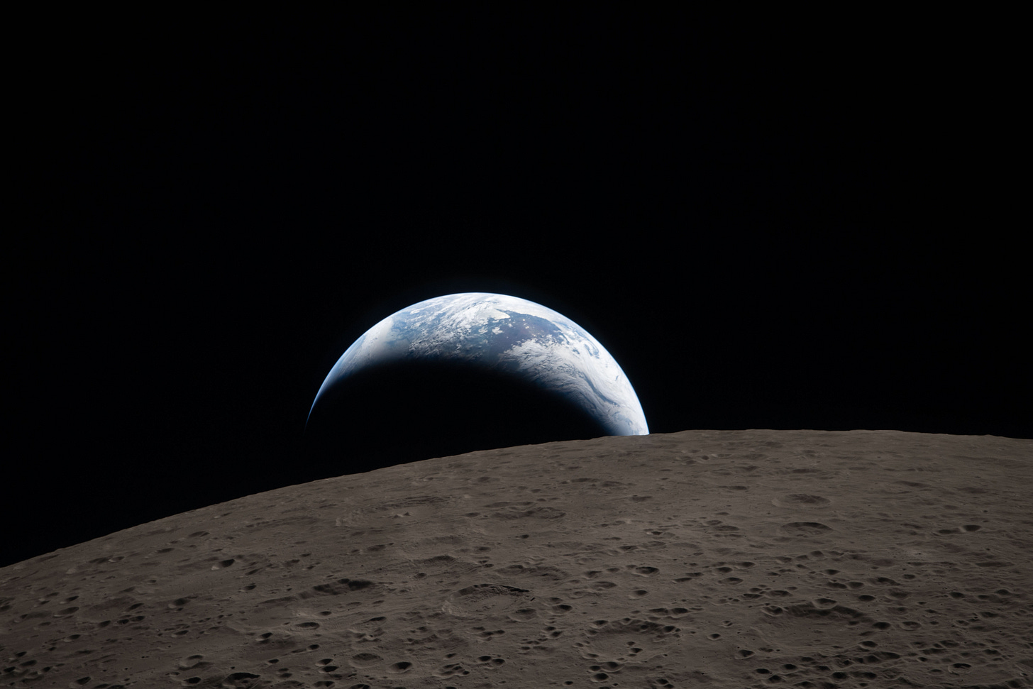 Earth setting behind the Moon’s horizon. Set against the blackness of space, the dark gray surface of the Moon arc across the image in the foreground. Beyond, a blue and white crescent Earth, oriented like a frown, fills the center of the image, with one tip disappearing beyond the lunar limb.