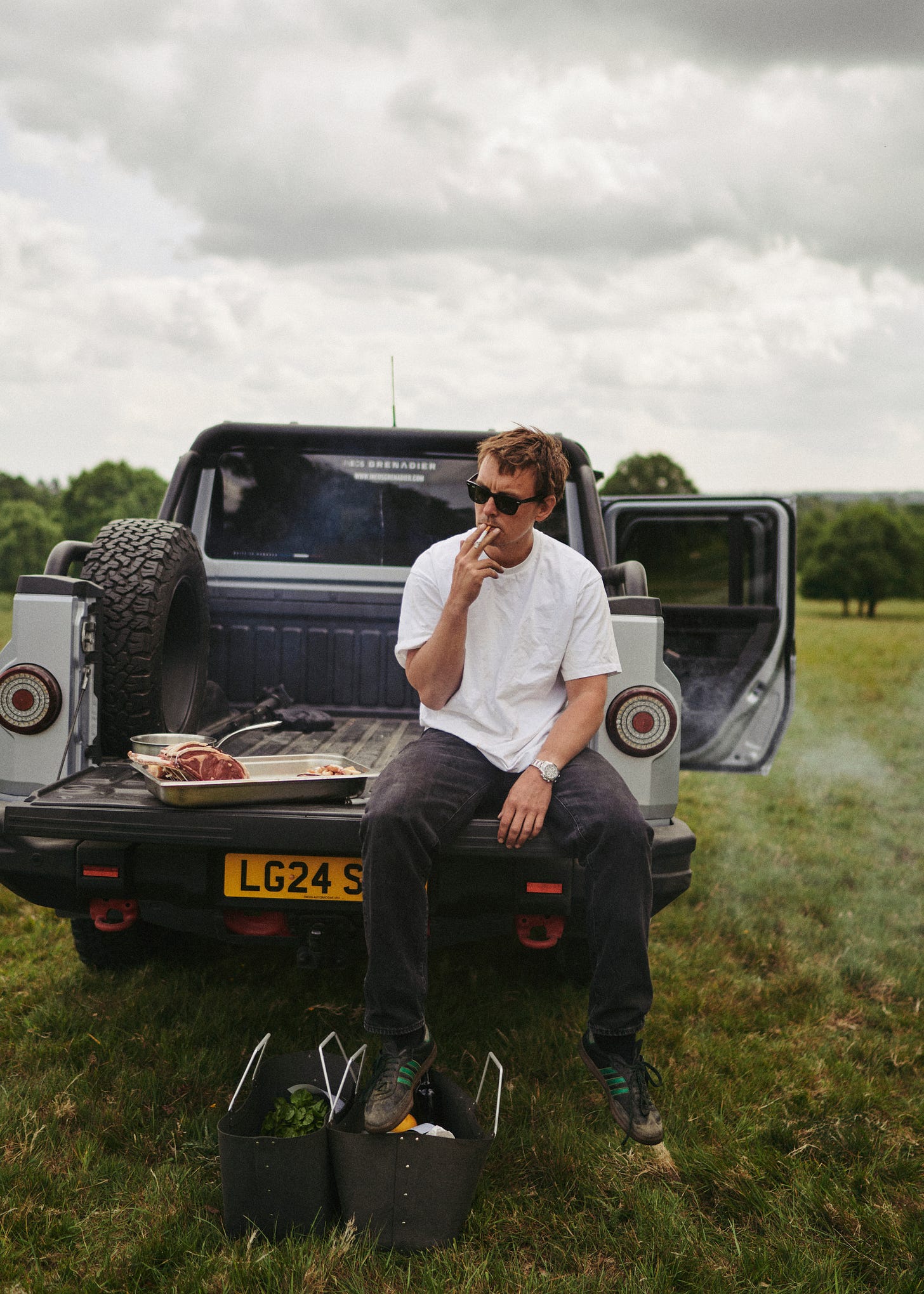 A man smoking a cigarette on the back of a truck outside.