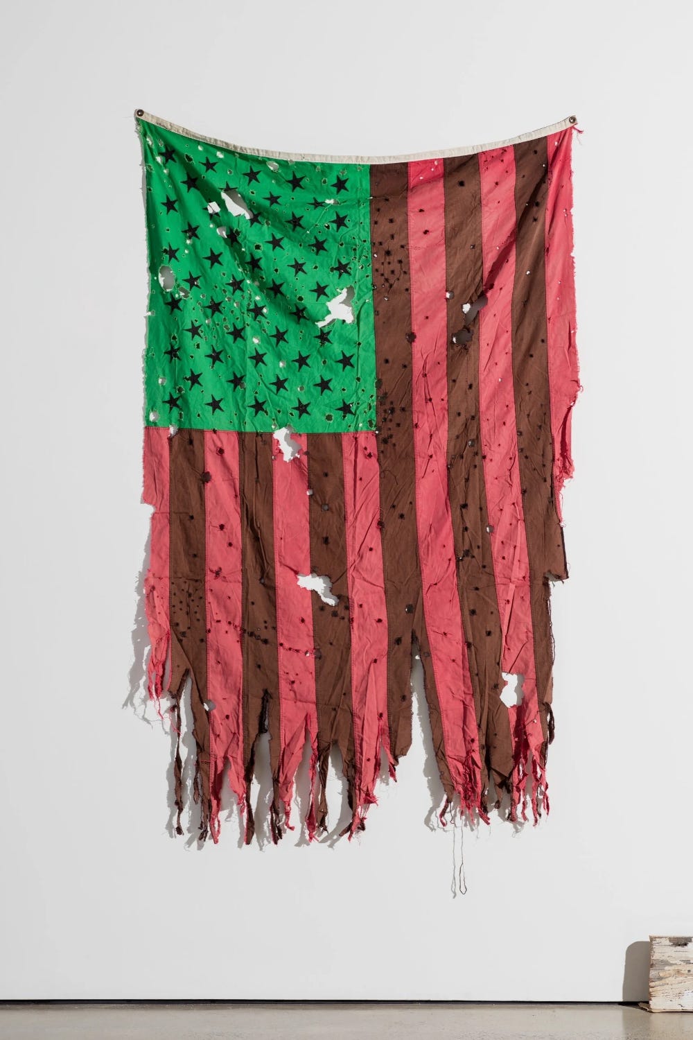 Artwork titled "Oh Say Can You See" by David Hammons shows a tattered Stars and Stripes flag peppered with bullet holes and other fabric damage. The stars are black on a green background, and the stripes are alternating red and brown. An Afrofuturist remix of a familiar symbol.
