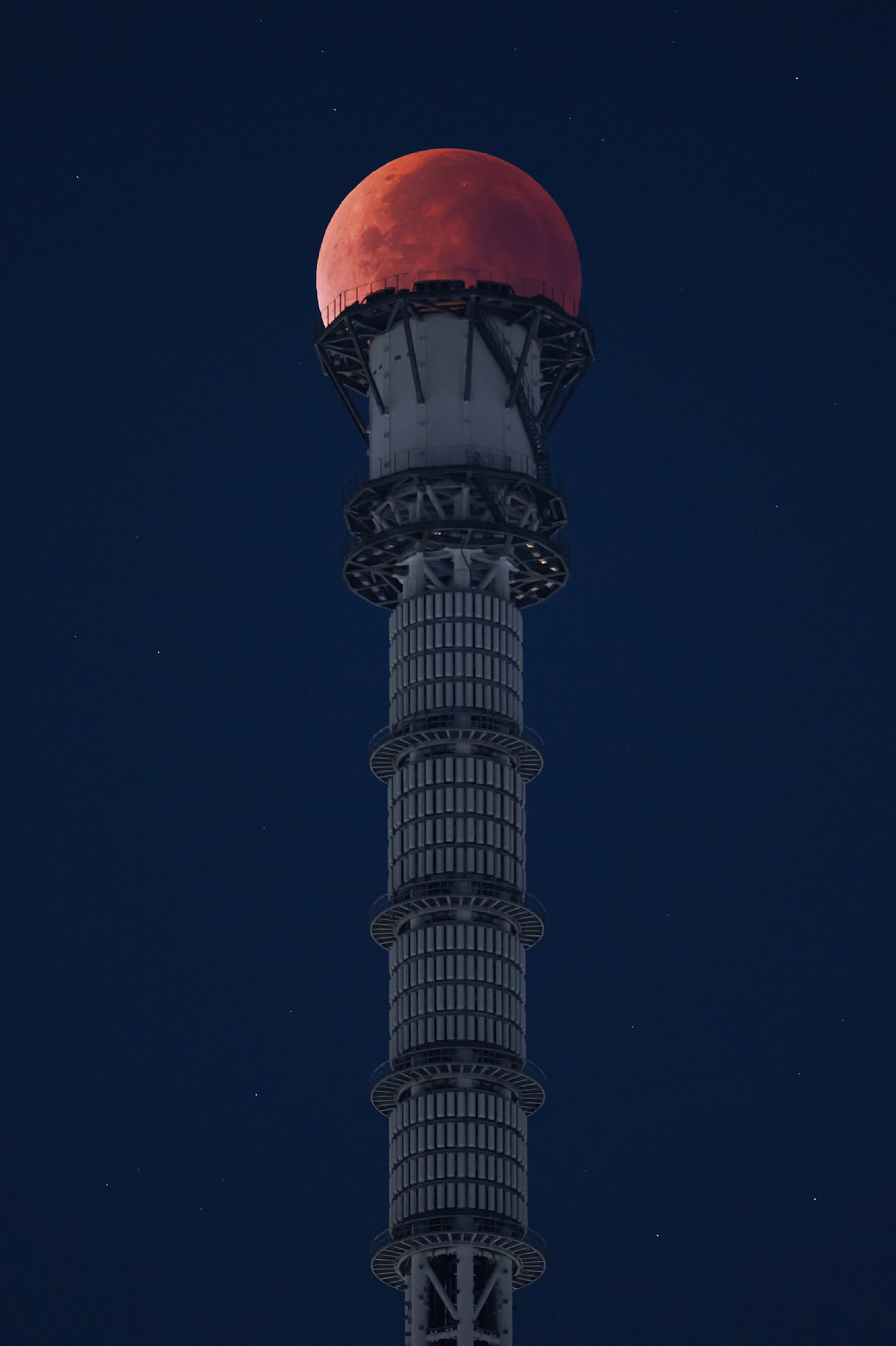 A red full moon is positioned atop a tower in Japan against a dark blue night sky. A red full moon is positioned atop a tower in Japan against a dark blue night sky.