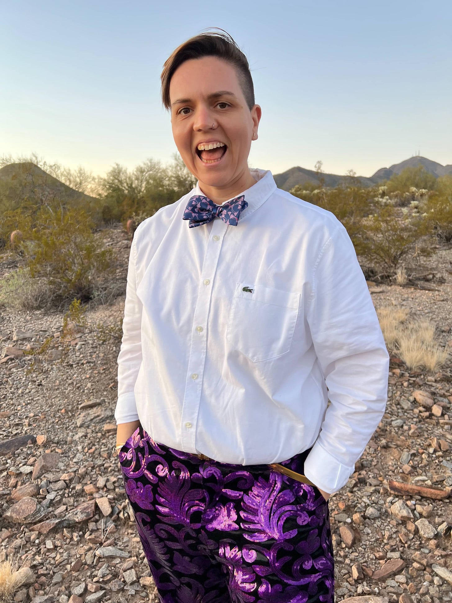 Dr. SG Gettman stands on desert landscape wearing long sleeve white button up, purple sequined pants, and a purple bowtie. Dr. gettman has short brown hair swept to the side. 
