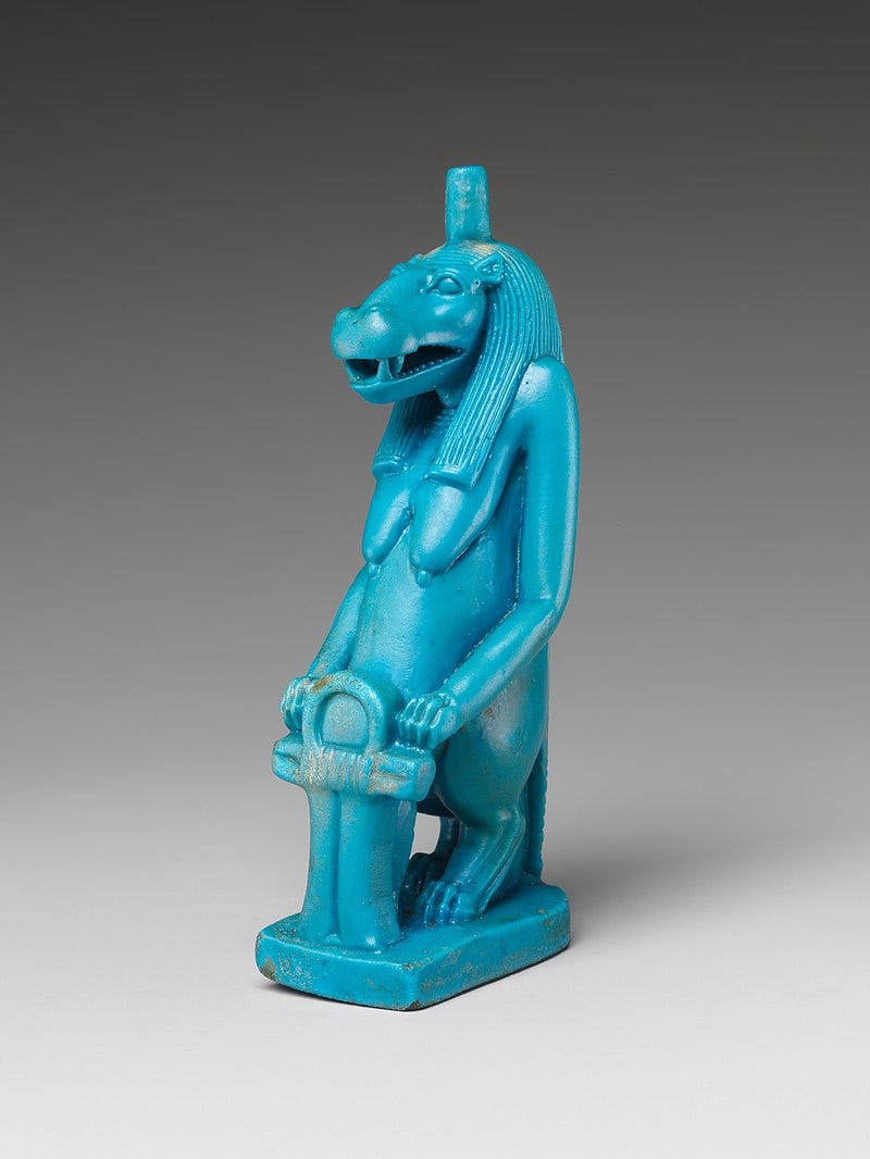 A turquoise bipedal hippo with large drooping breasts and braided hair that falls over its shoulders stands holding a key of life. A turquoise bipedal hippo with large drooping breasts and braided hair that falls over its shoulders stands holding a key of life.
