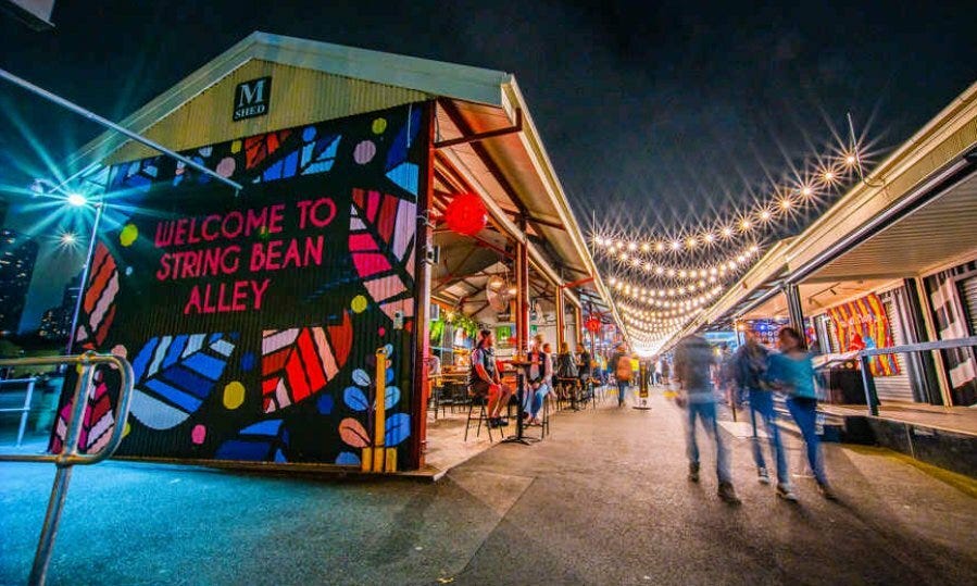 Nighttime view of String Bean Alley at Queen Victoria Market in Melbourne, featuring colourful mural signage, festoon lighting, and people walking past small shops and eateries.