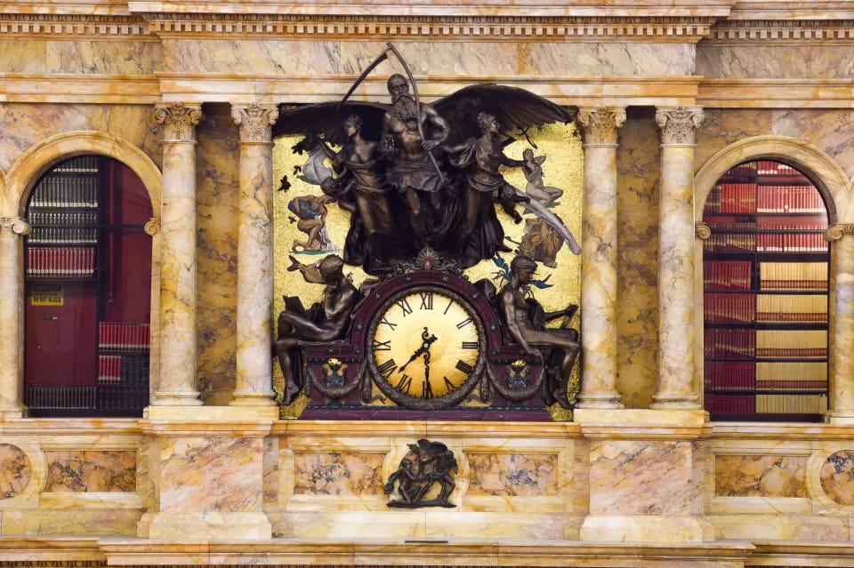 Ornate bronze clock with allegorical sculptures of Father Time and figures, set into a marble wall flanked by library bookshelves. Ornate bronze clock with allegorical sculptures of Father Time and figures, set into a marble wall flanked by library bookshelves.