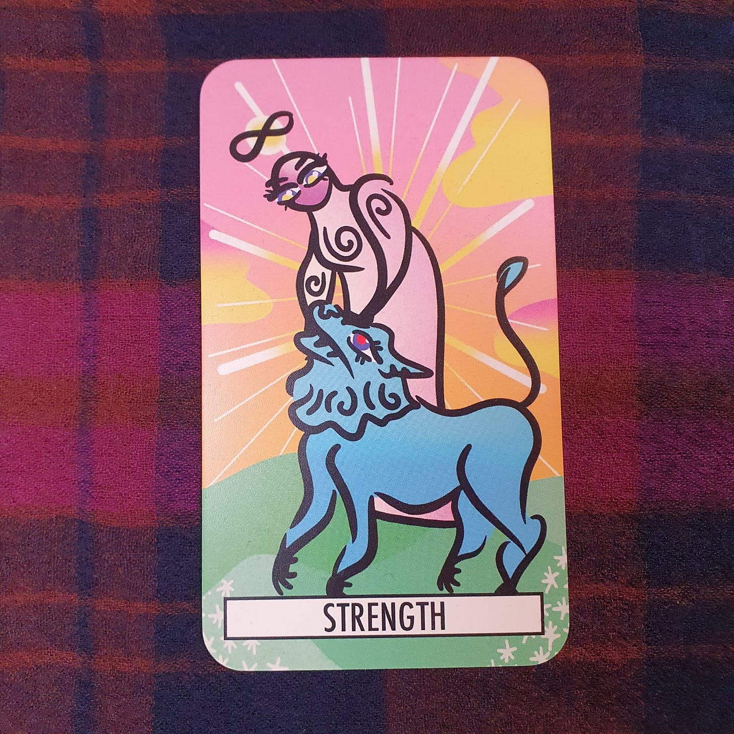 Strength card from The Theatre Deli Tarot, showing a stylised figure with an infinity symbol above their head and a blue lion beneath a radiant pastel sky. Tarot card drawn during Amy Twigg’s Tarot DMs interview to discuss resilience, recovery, courage and quiet inner strength. Strength card from The Theatre Deli Tarot, showing a stylised figure with an infinity symbol above their head and a blue lion beneath a radiant pastel sky. Tarot card drawn during Amy Twigg’s Tarot DMs interview to discuss resilience, recovery, courage and quiet inner strength.