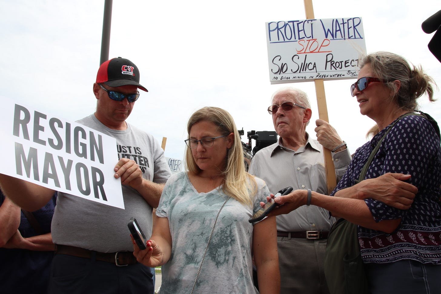 Three people stand around a woman holding her phone out. The others hold signs reading "Resign mayor" and "Protect water." Three people stand around a woman holding her phone out. The others hold signs reading "Resign mayor" and "Protect water."