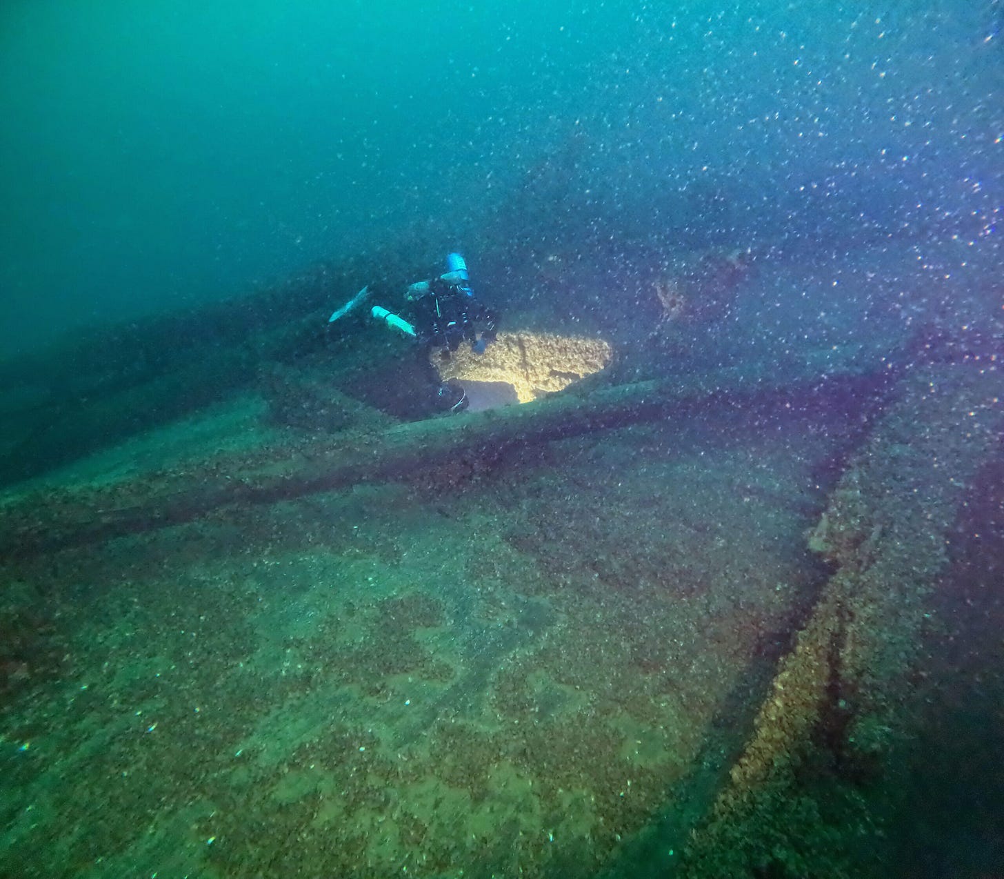 A scuba diver explores the interior of a sunken shipwreck on the ocean floor, surrounded by scattered debris and low visibility.