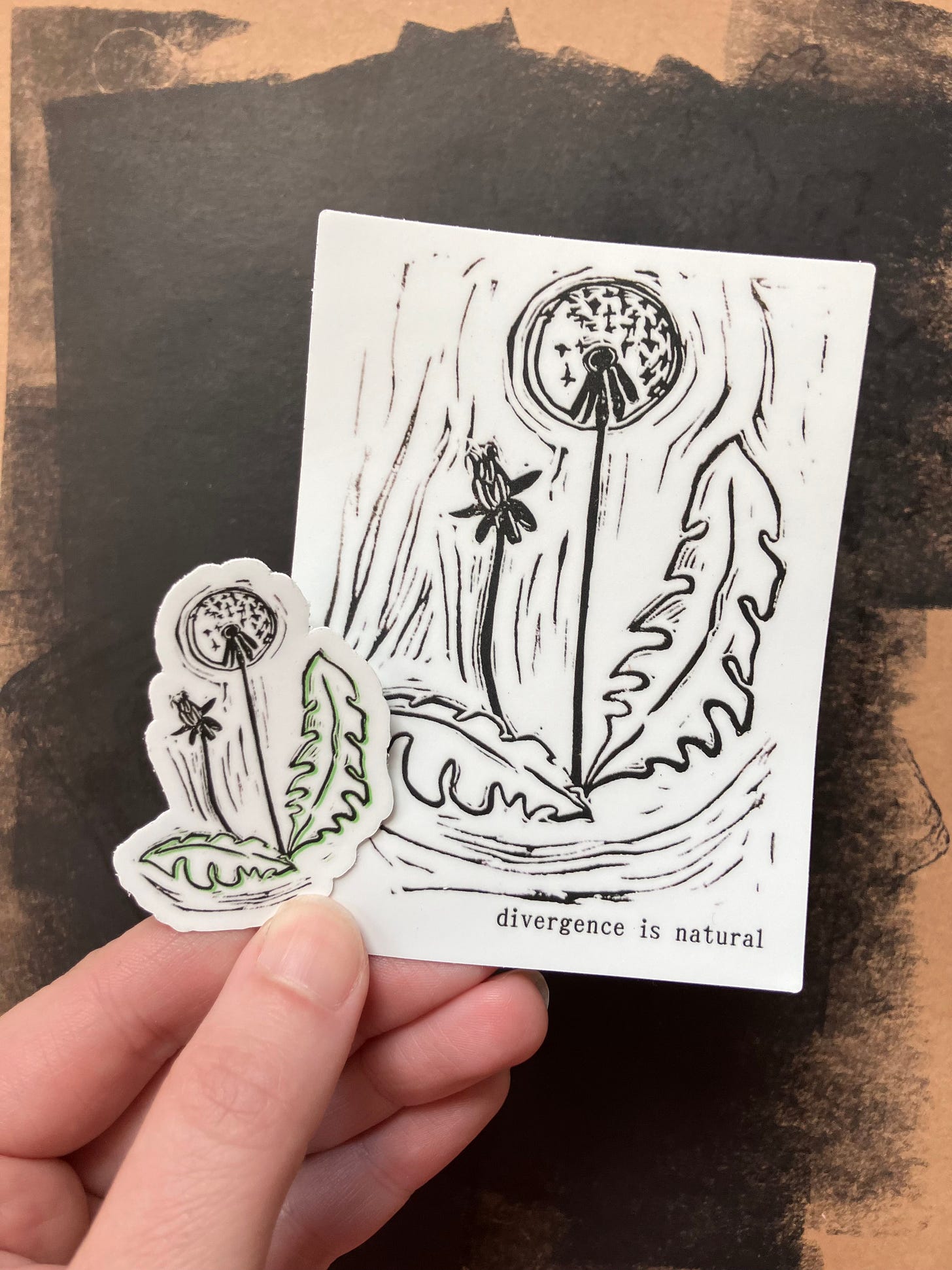 A photo of two stickers on a black background. One sticker is a large dandelion with the text, "Divergence is natural." The other sticker is a small dandelion with a green outline. A photo of two stickers on a black background. One sticker is a large dandelion with the text, "Divergence is natural." The other sticker is a small dandelion with a green outline.