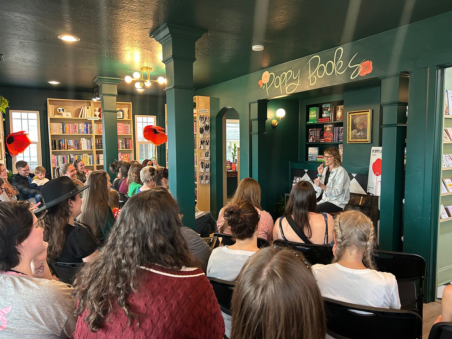 A photo of Sooz in front of a cute bookshop filled with people. she's grinning and talking into a mic while everyone watches on, smiling too.