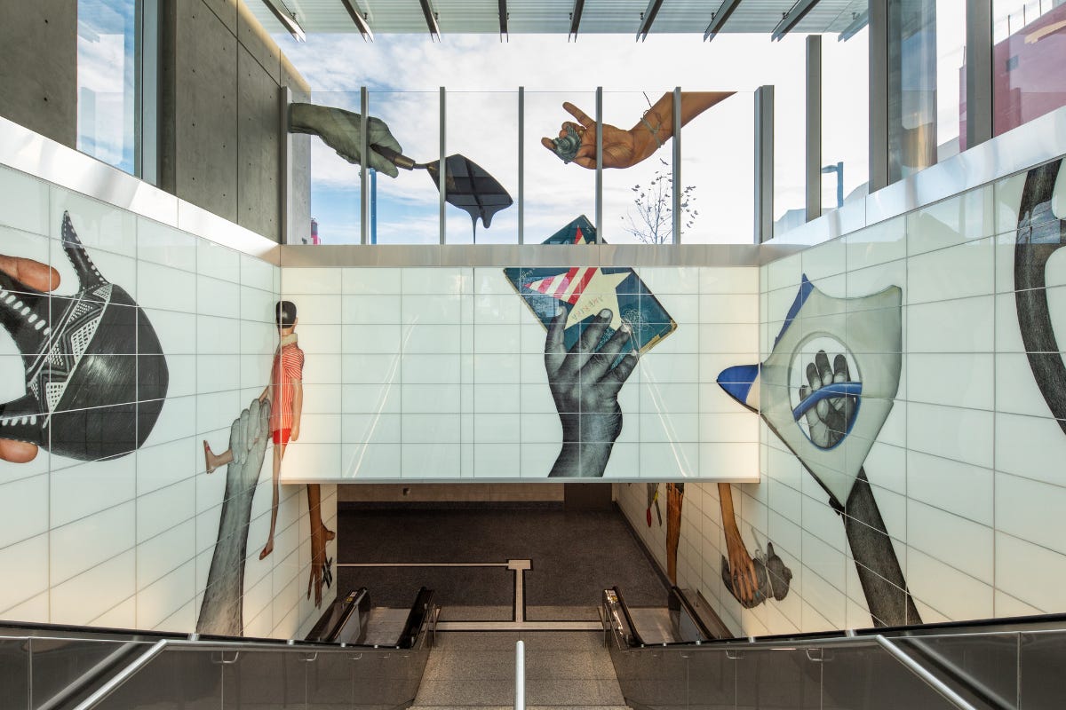 A large-scale station artwork showing oversized hands holding objects across tiled walls above an escalator, with images of tools, vessels, and figures spread across the station architecture. A large-scale station artwork showing oversized hands holding objects across tiled walls above an escalator, with images of tools, vessels, and figures spread across the station architecture.