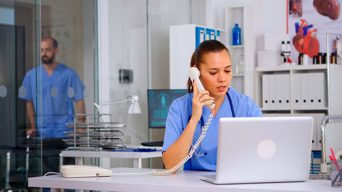 Infermiere al telefono seduta a una scrivania in uno studio medico moderno, con un laptop davanti e documenti clinici. Sullo sfondo, un collega in camice azzurro visibile dietro una parete in vetro.