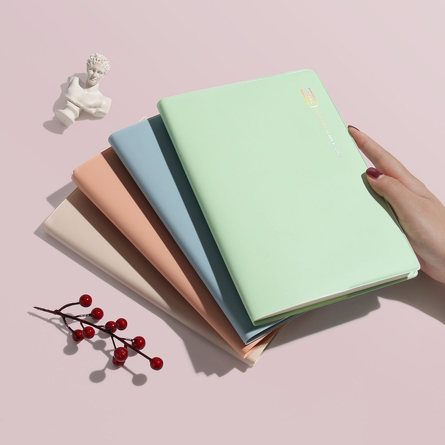 A hand resting on four stacked journals in colours green, blue, pink, and white - on a pastel pink background A hand resting on four stacked journals in colours green, blue, pink, and white - on a pastel pink background