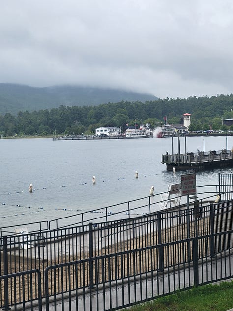 A collection of 9 images depicting: the shoreline of Lake George including a lighthouse and steamboat; a beautiful sunset with glowing pink clouds over the lake, being photographed from a deck behind a woman with long, wet hair who is photographing the same clouds; the lake on a sunny day with water sparkling in front of low mountains; a duck looking at the camera from a sandy beach; a young teenage boy sitting at a table with a small town street in the background; the same teenage boy wearing a wet swimsuit, sitting astride a bear statue at an indoor water park; the same teenage boy sitting with his feet up in his chair on the deck by the lake, wearing a coat and headphones as he stairs at his phone; a street view of the small downtown area with mountains rising in the background; pink, fluffy, glowing clouds over the lake at sunset.