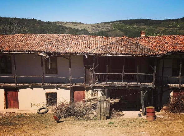 una fattoria di Arbanasi, in Bulgaria: un edificio lungo su due piani, con tetto in tegole e balcone di legno, sullo sfondo colline e cielo azzurro e in primo piano galline libere nell'aia
