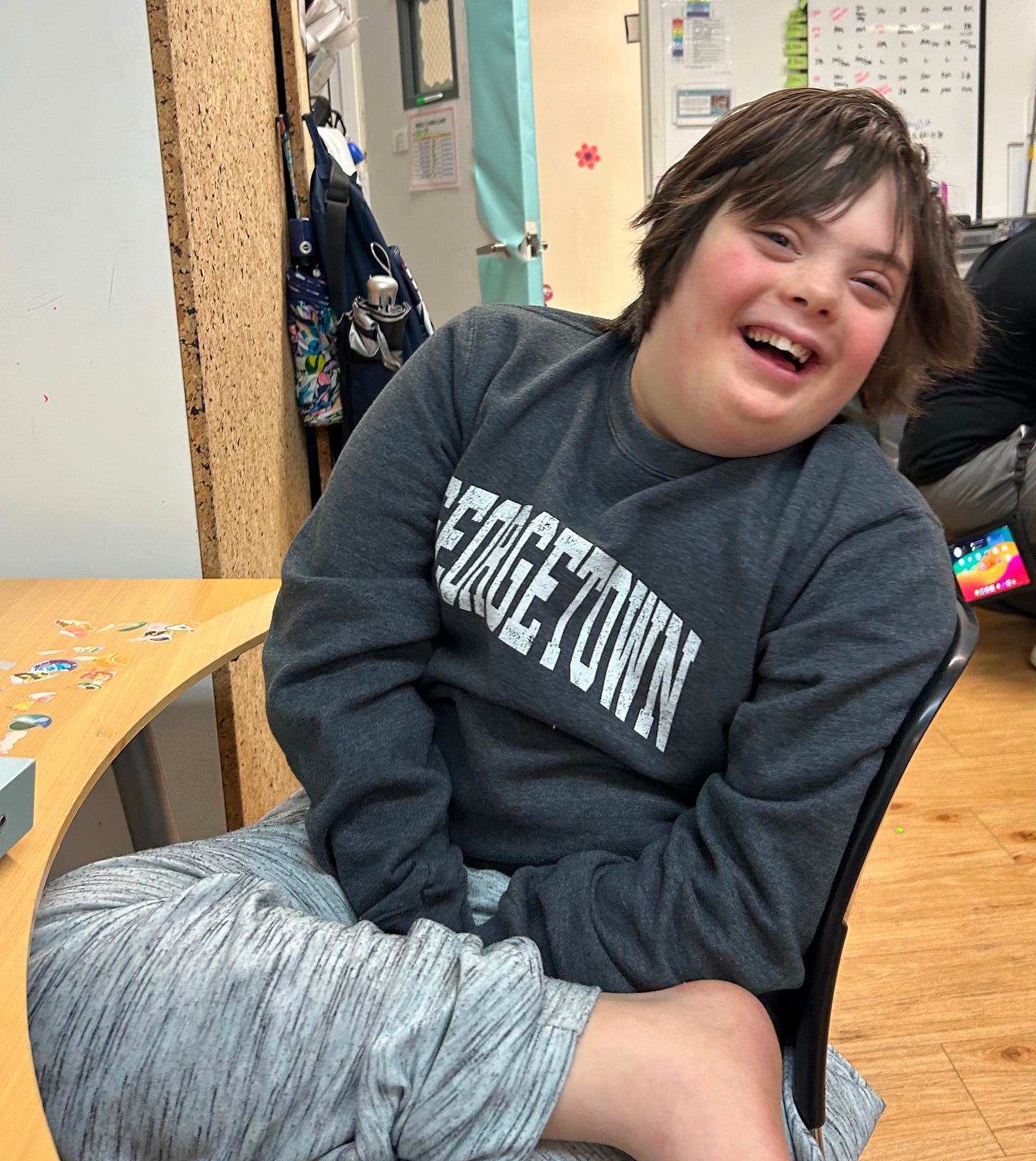 A white boy with Down syndrome laughing, seated at a desk. 