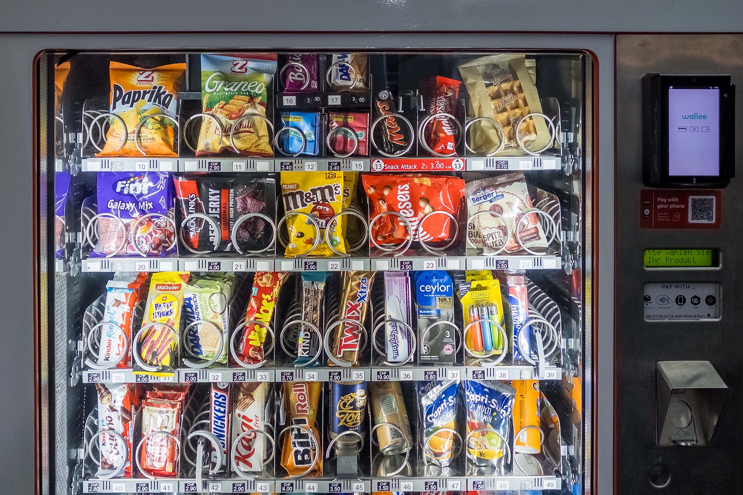Das Foto zeigt die Frontansicht eines typischen Verkaufsautomaten, wie er in Unternehmen, Schulen oder Bahnhöfen zu finden ist. Der Automat ist gefüllt mit einer Vielzahl von Snacks, die überwiegend als ungesund gelten, wie Chips, Schokoriegel (Snickers, Twix, Mars), Gummibärchen, M&M's und andere Süssigkeiten. Nur wenige gesündere Alternativen wie Müsliriegel und Trockenfrüchte. Zusätzlich sind unten Capri-Sonne und oben vereinzelt salzige Snacks wie Paprika-Chips sichtbar. Rechts am Automaten befinden sich ein Kartenzahlungsterminal sowie ein QR-Code für mobiles Bezahlen.