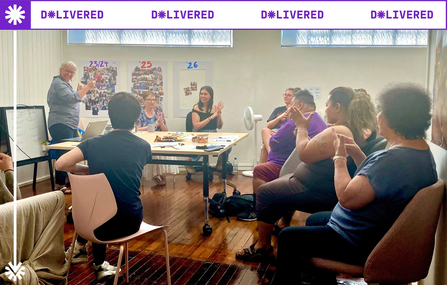 a group of adults sitting around a table practising NZSL signs. Monica Leach facilitates the session in The D*List community space
