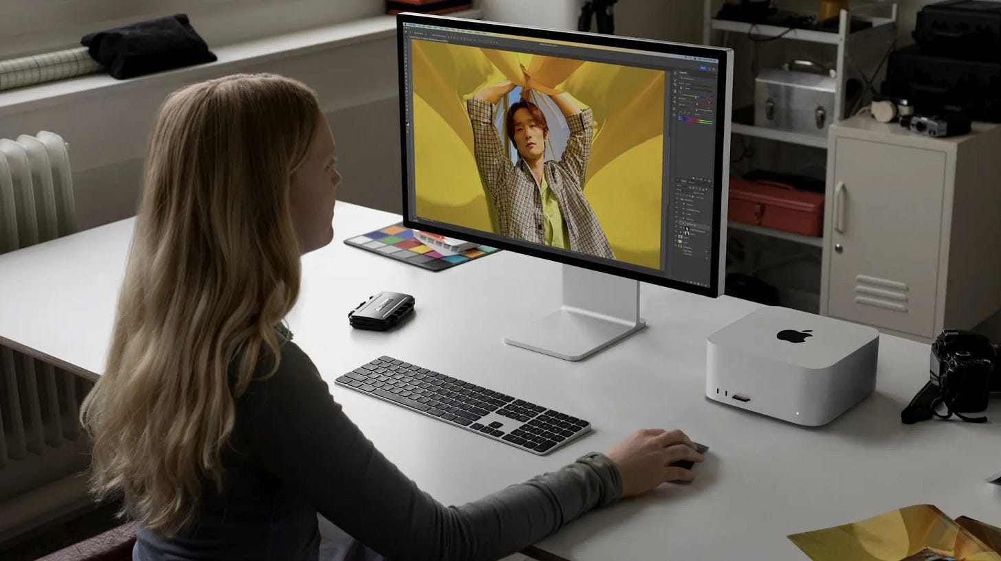 A photo editing workstation showing a person with blonde hair working on a large professional monitor displaying a portrait with yellow background. The setup includes an Apple Mac Mini, color calibration card, and clean white desk in a photography studio environment.