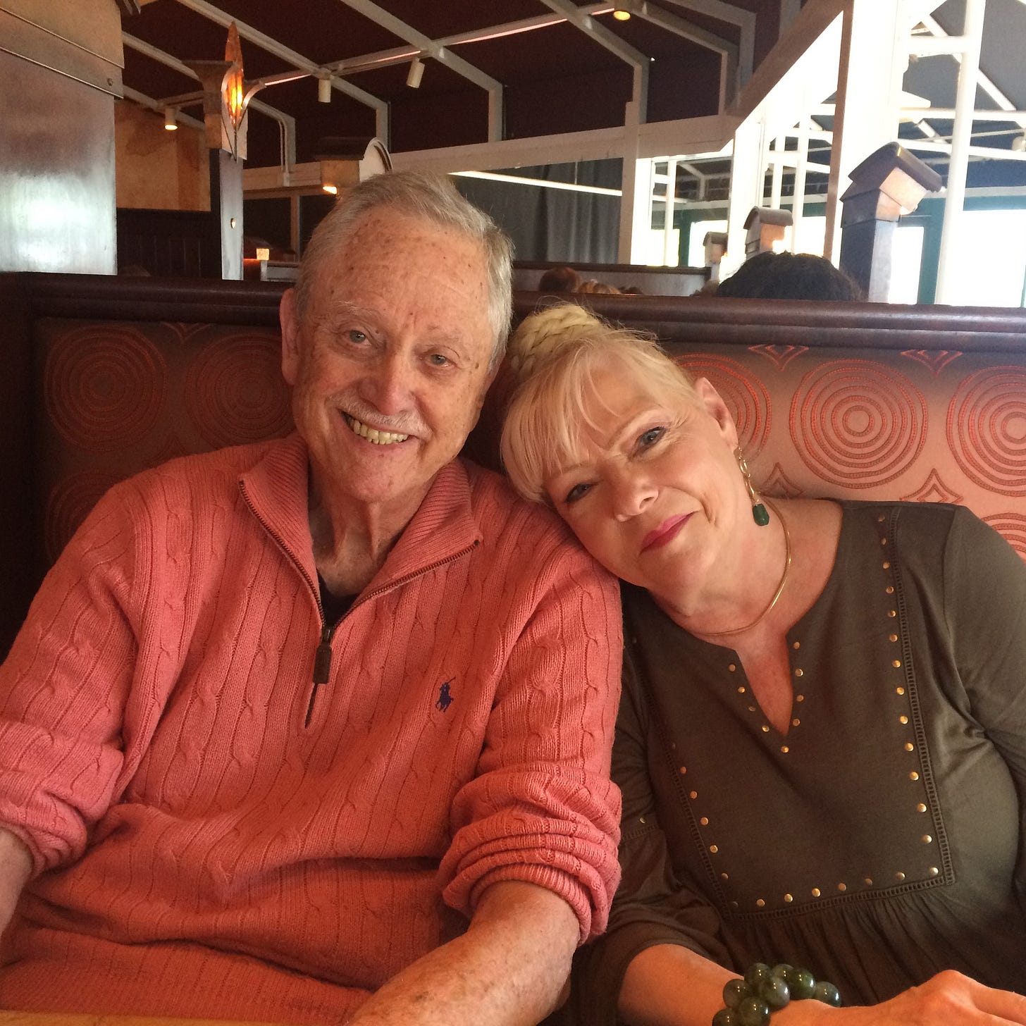 A man in a salmon colored sweater is seated beside a blonde woman, resting her head on his shoulders A man in a salmon colored sweater is seated beside a blonde woman, resting her head on his shoulders