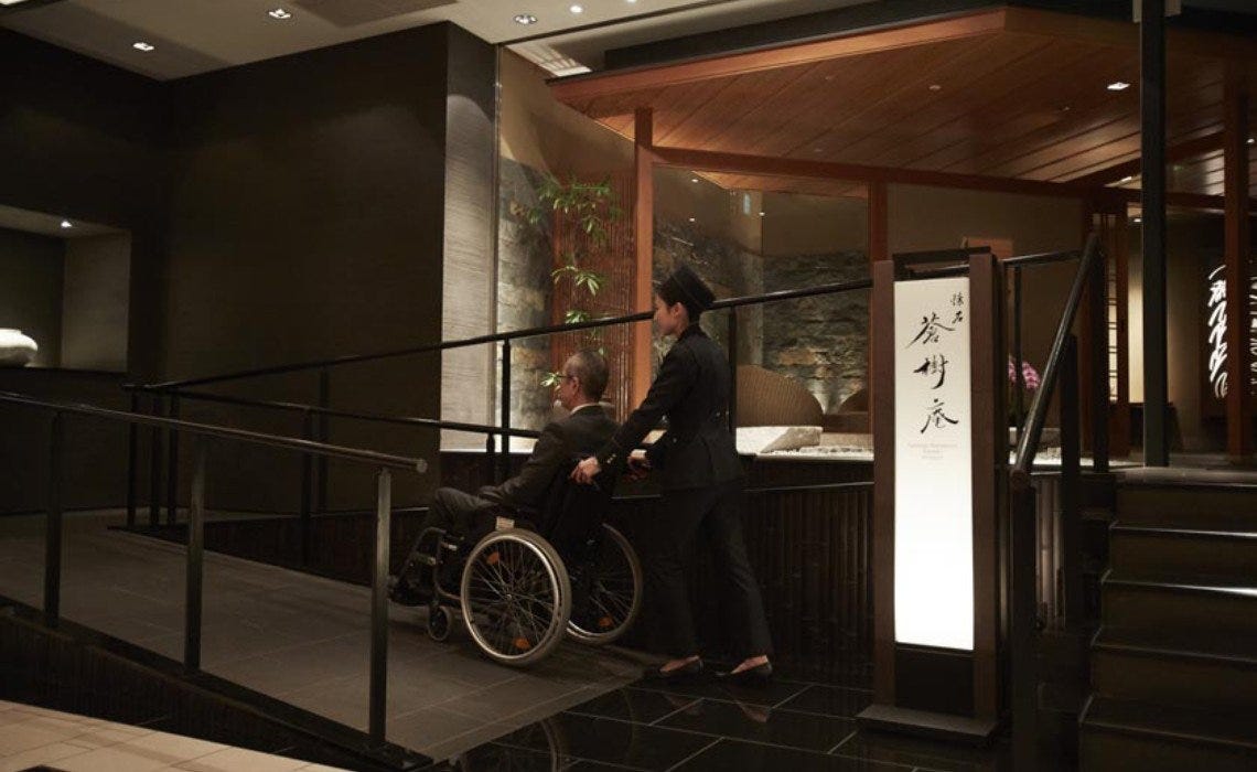 A hotel staff member assists a guest using a wheelchair up an indoor ramp toward the entrance of a modern Japanese restaurant with wooden accents and soft lighting.