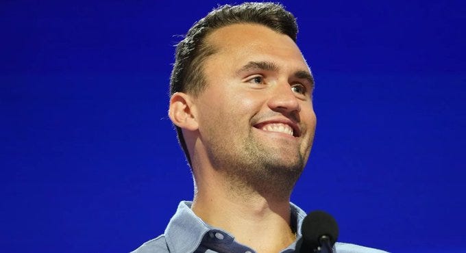Charlie Kirk standing on a stage, wearing a light blue shirt, speaking into a microphone against a blue background.