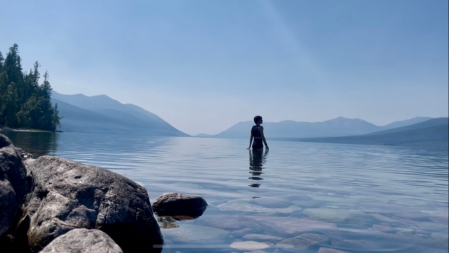 A photograph of Lyss standing in a crystal clear lake against a background of blue mountains and evergreen trees.
