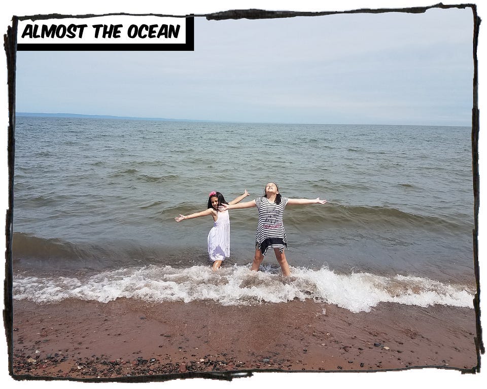 Two girls standing in the waves of Lake Superior with their arms spread wide. The caption reads, “Almost the ocean.”