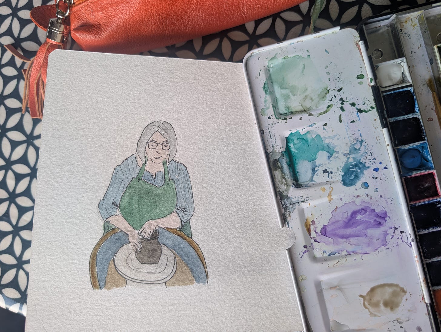 A loose pen-and-watercolour sketchbook illustration of Andrea sitting at a potter's wheel, shaping a lump of clay with both hands while wearing an apron. A paint palette and brushes rest in the foreground, emphasising the creative process. The gentle illustration accompanies a reflection on how pressure and preparation can shape both clay and our own lives. A loose pen-and-watercolour sketchbook illustration of Andrea sitting at a potter's wheel, shaping a lump of clay with both hands while wearing an apron. A paint palette and brushes rest in the foreground, emphasising the creative process. The gentle illustration accompanies a reflection on how pressure and preparation can shape both clay and our own lives.