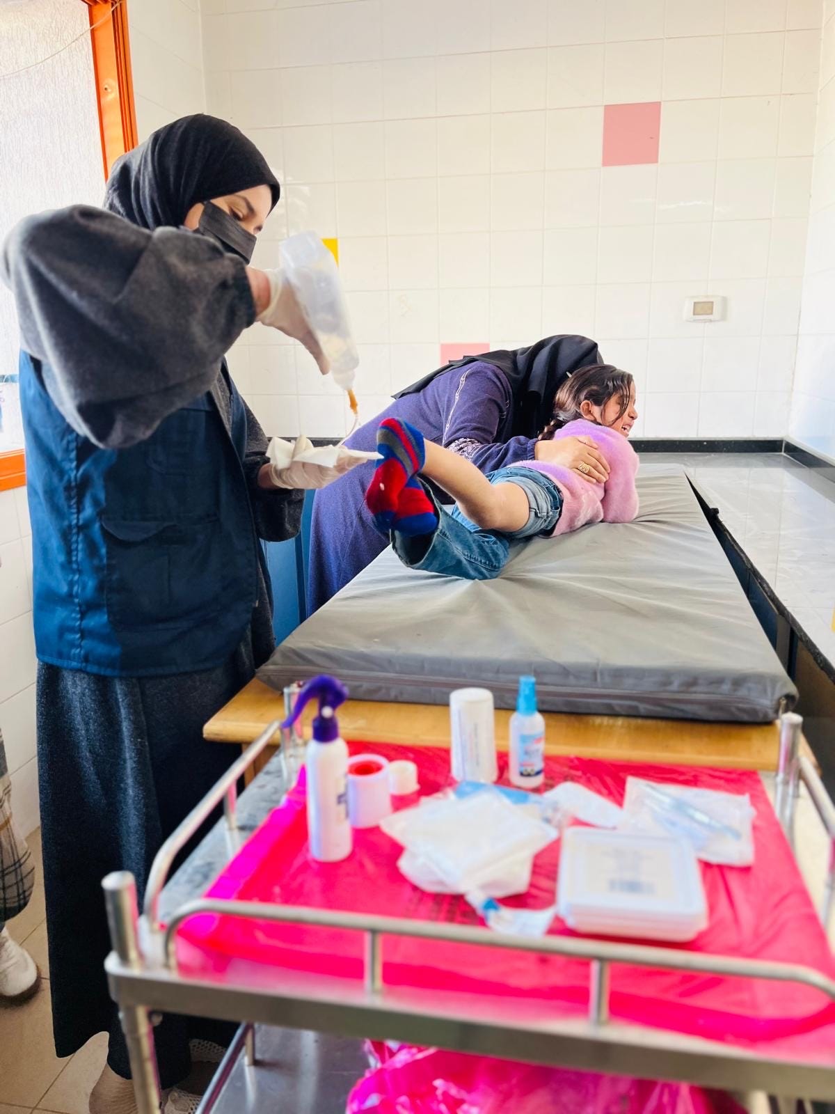 A healthcare worker wearing a hijab and face mask is preparing medical supplies while another woman holds a young child on a medical examination table. The child appears distressed. Medical supplies, including bandages, spray bottles, and ointments, are organized on a cart in the foreground. The room has tiled walls and bright lighting.