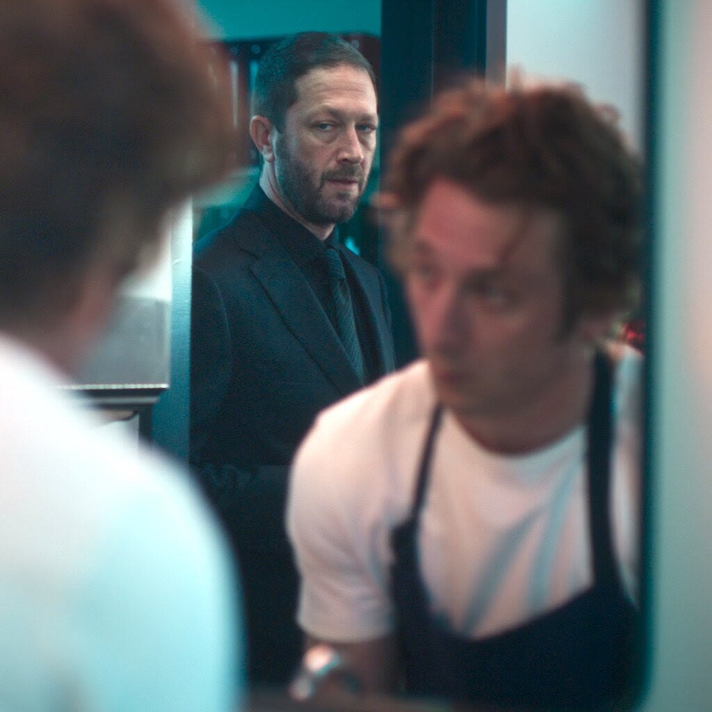 In a scene from “The Bear,” Ebon Moss-Bachrach, in a dark suit, looks into a mirror at Jeremy Allen White, who is wearing an apron.