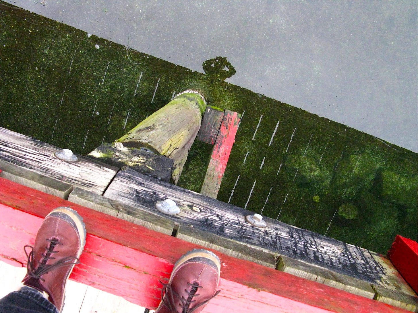 Foreground shoes on edge of dock, reflection in water of person leaning over dock taking photo