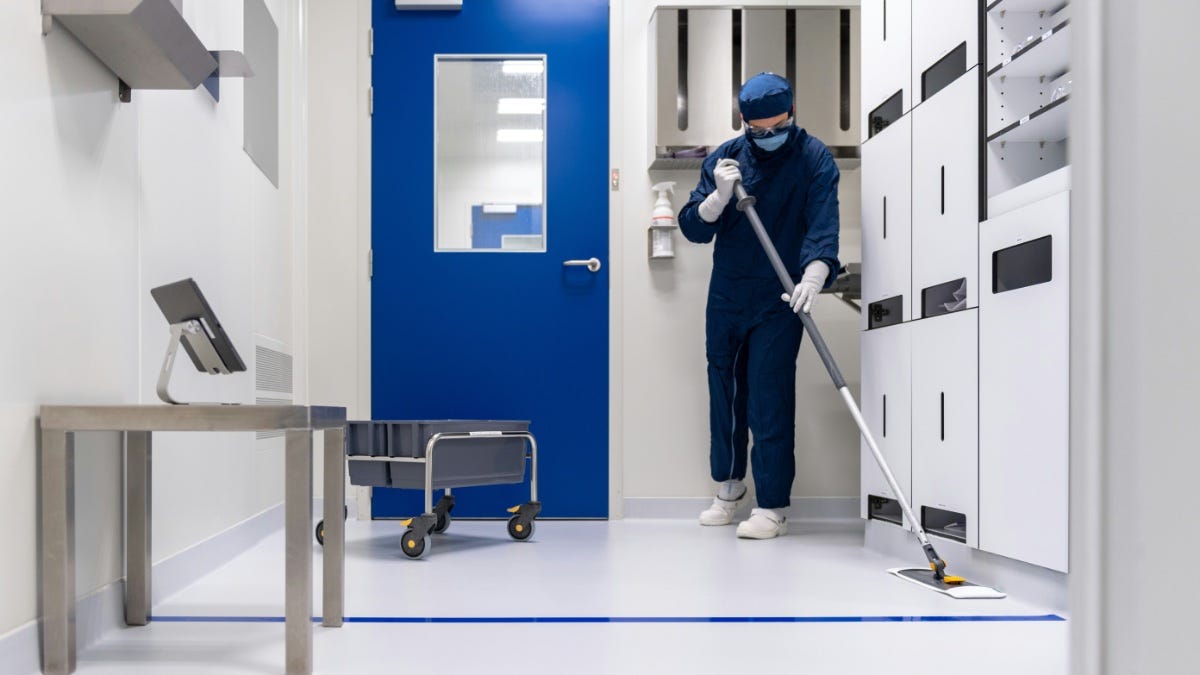 A person in protective clothing mops the floor of a cleanroom with blue and white walls, a cart, and a mounted monitor nearby. A person in protective clothing mops the floor of a cleanroom with blue and white walls, a cart, and a mounted monitor nearby.