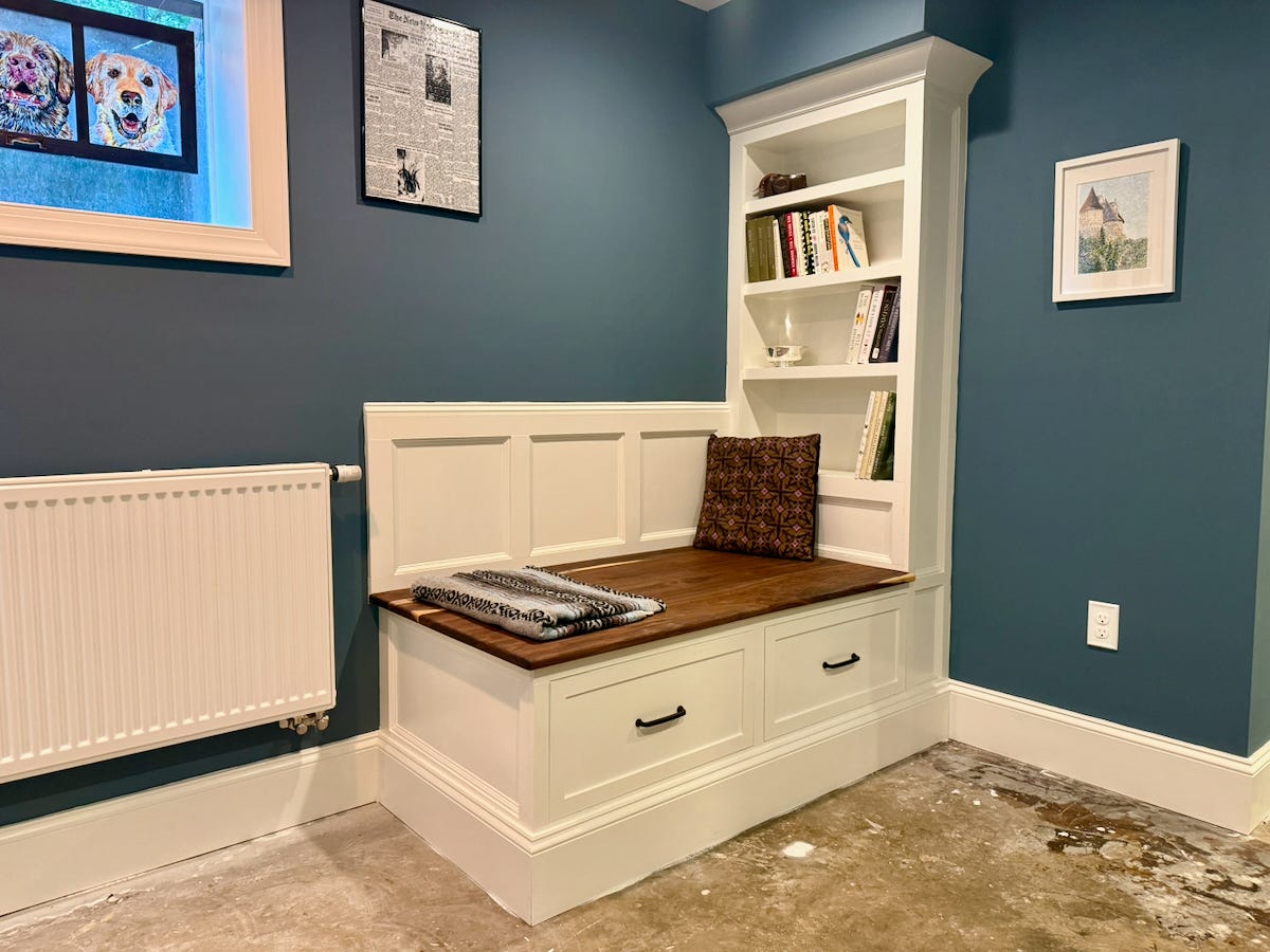 A custom built-in reading bench and bookshelf installed in a basement nook. The bench features a solid walnut seat above two large storage drawers painted white with black pulls. Matching white paneled wainscoting rises behind the bench to form a backrest. A tall built-in bookshelf with crown molding stands to the right, filled with a small collection of books and decorative objects. The walls are painted a deep blue, contrasting with the white trim, while a white radiator sits to the left beneath a small window with stained-glass dog artwork. The unfinished concrete floor in the foreground highlights the newly completed built-in before the room’s flooring has been installed.