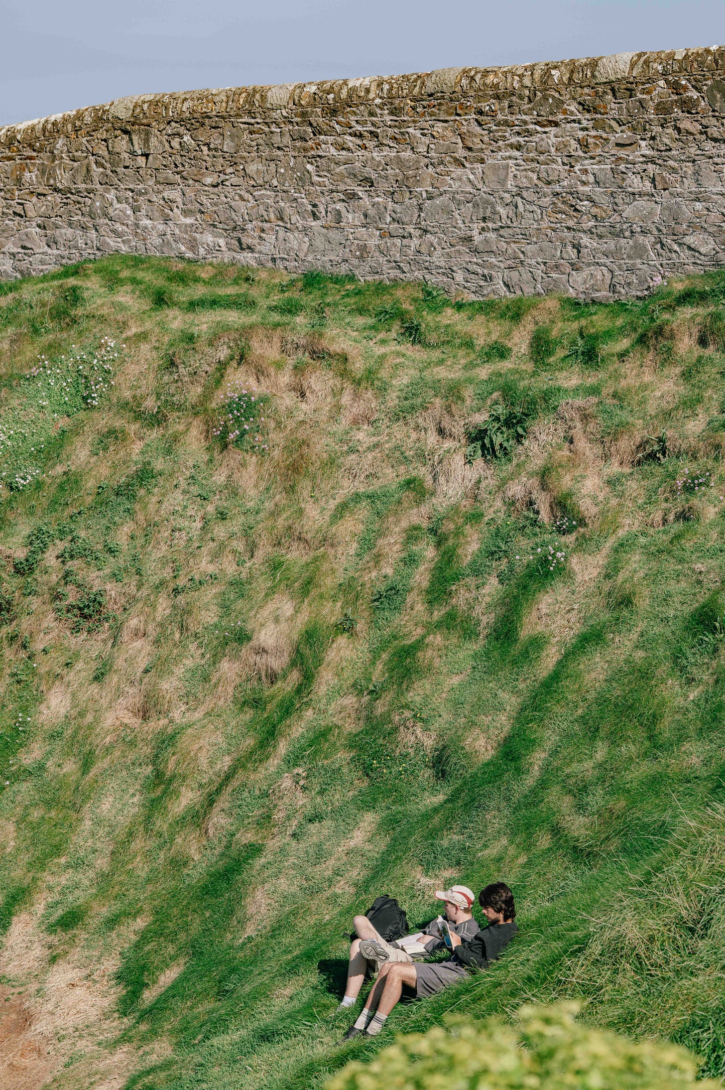Two people rest on the sloped grassy cliffs below a stone wall, the texture of the land patterned with brown and green patches. Two people rest on the sloped grassy cliffs below a stone wall, the texture of the land patterned with brown and green patches.