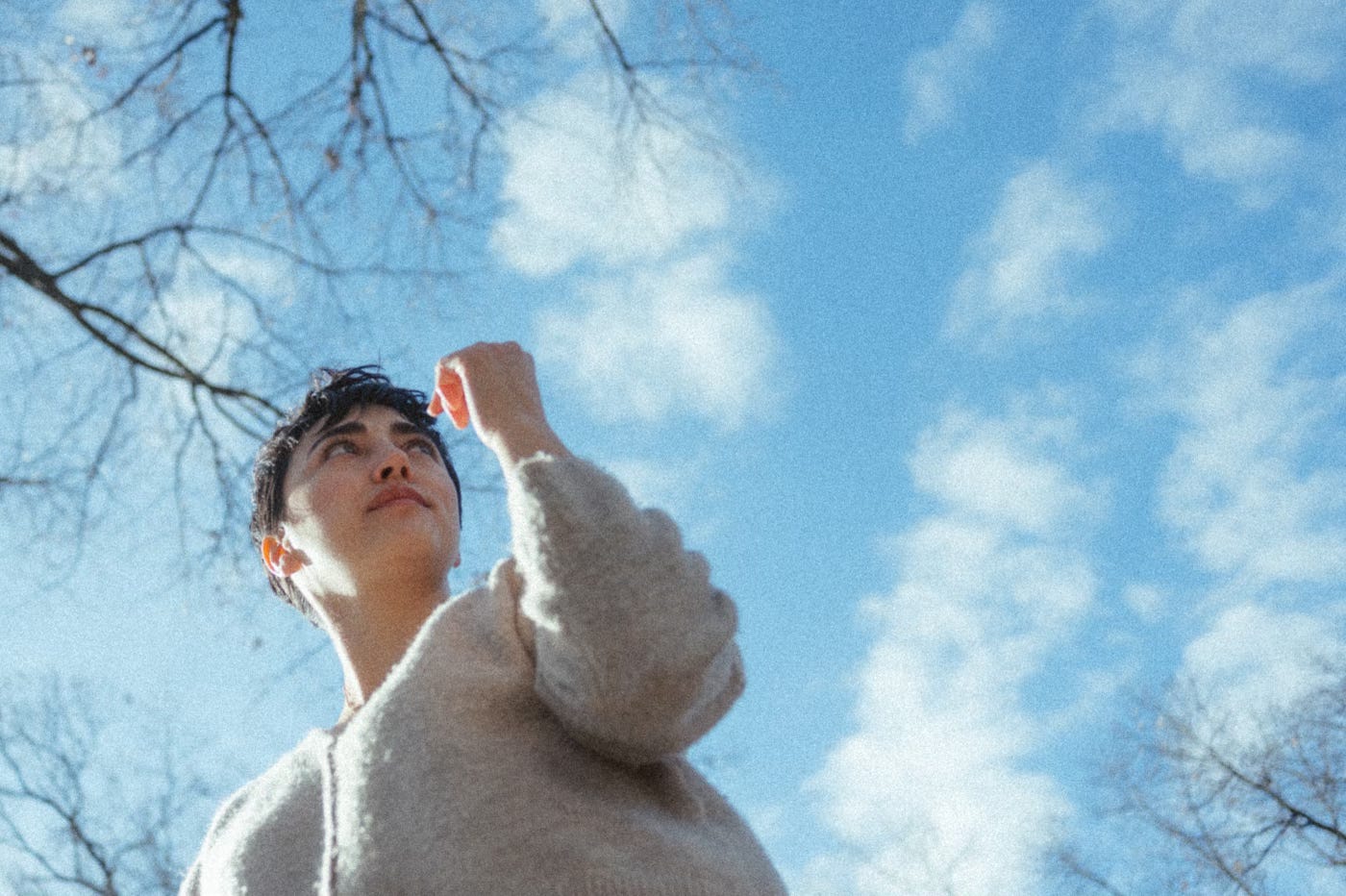 Alex Blue, who has short dark hair and tan skin, stands off to the left center. The photo is taken from below and the blue sky and some bare treetops can be seen above her.