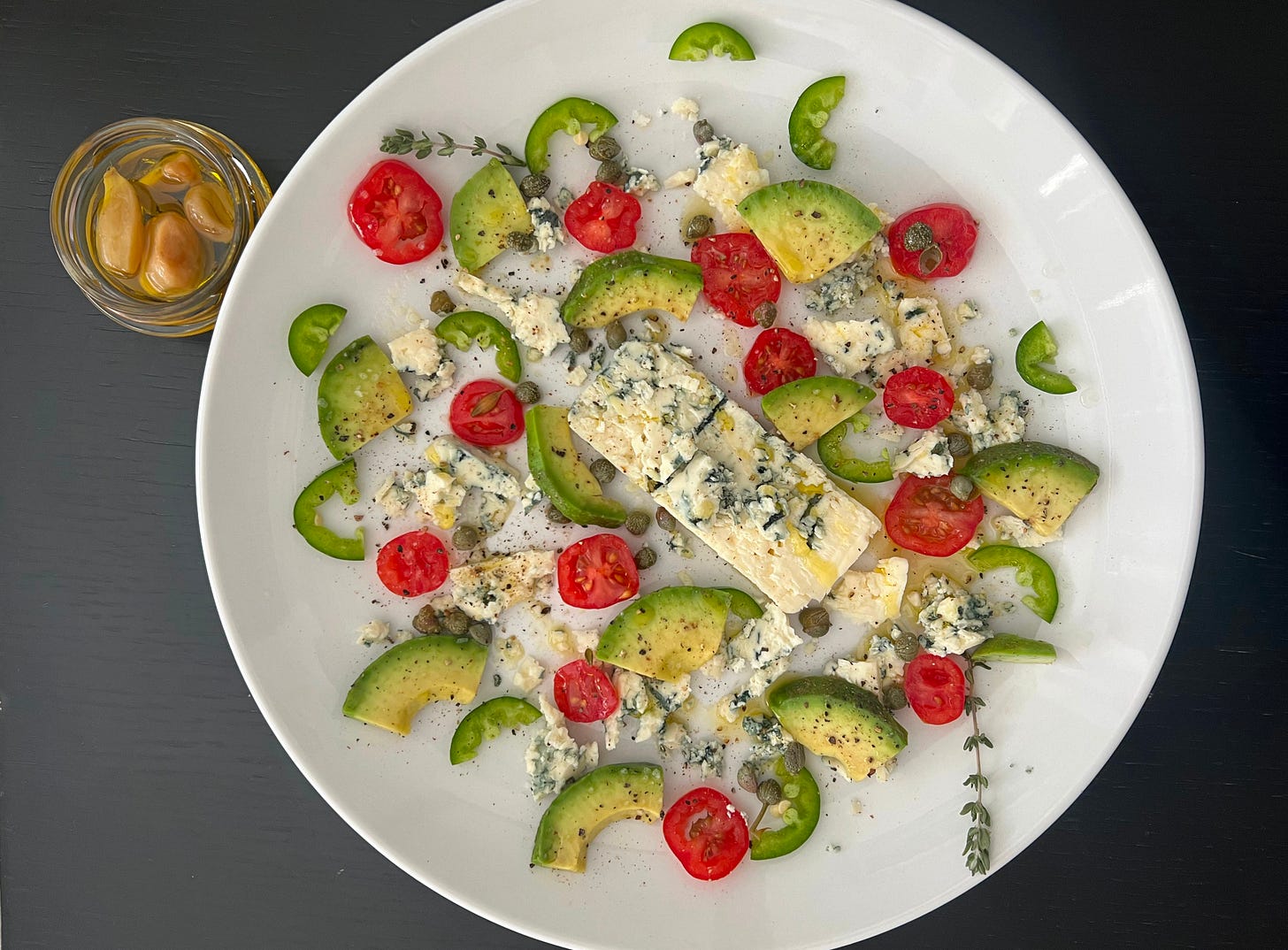 A vibrant platter featuring creamy avocado slices, chunks of blue cheese, and fresh cherry tomatoes arranged alongside capers and green bell peppers, all drizzled with olive oil and a touch of cracked pepper. A small glass bowl with confit garlic cloves sits beside the plate. The image embodies the essence of Mediterranean-inspired cuisine for Foodie Disorder, highlighting tomatoes' versatility. Presented by Liat Portal for The Liat Show. A vibrant platter featuring creamy avocado slices, chunks of blue cheese, and fresh cherry tomatoes arranged alongside capers and green bell peppers, all drizzled with olive oil and a touch of cracked pepper. A small glass bowl with confit garlic cloves sits beside the plate. The image embodies the essence of Mediterranean-inspired cuisine for Foodie Disorder, highlighting tomatoes' versatility. Presented by Liat Portal for The Liat Show.
