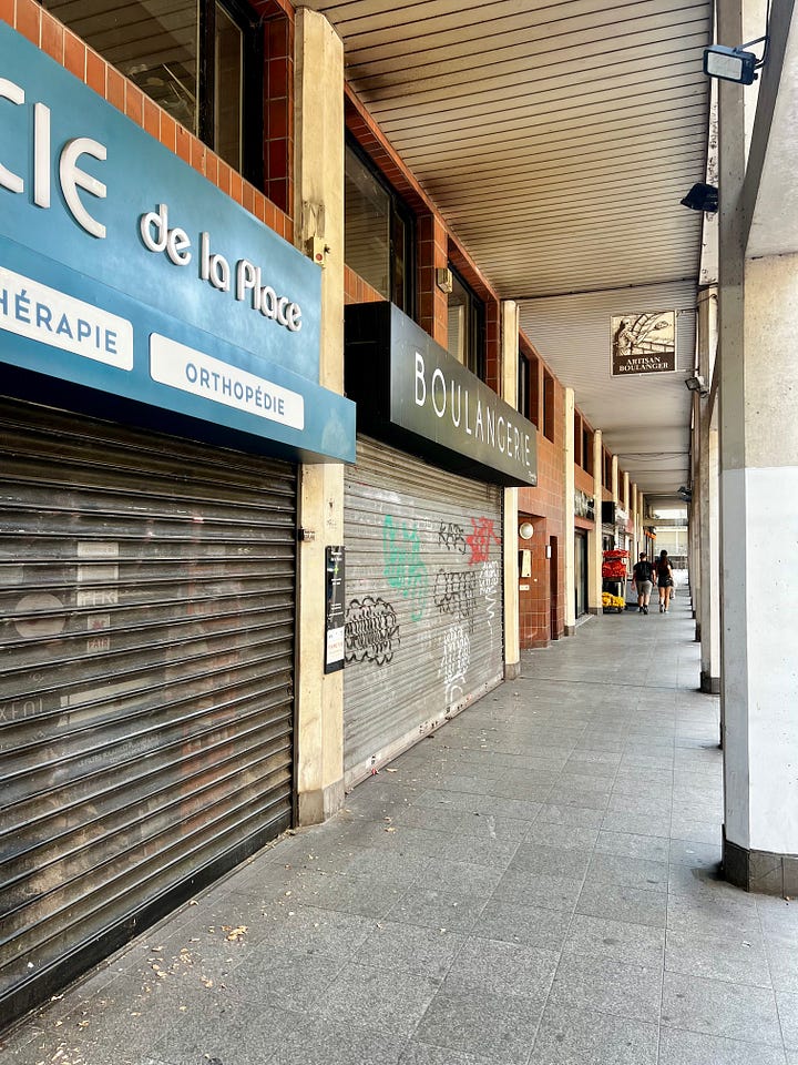 (emptEmpty café chairs arranged outside the Daumesnil metro station entrance on a quiet afternoon, with trees casting shadows on the pavement. Row of shuttered storefronts along an empty Parisian street, with closed metal security gates pulled down over shop windows.