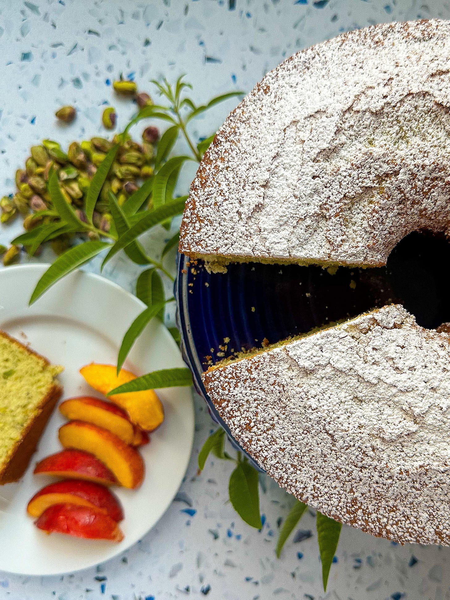 Photo of Lemon verbeana cake on a cake plate sitting next to a plate with a slice of cake and sliced peaches.