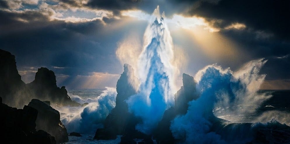 A dramatic coastal scene featuring dark, craggy rock formations on the left and right, framing a powerful ocean surge in the center. Waves crash against the rocks with white foam, while in the middle, a towering, soft plume of water erupts upwards like a geyser, illuminated by a bright, almost ethereal light from above. The sky is dark and cloudy on the edges, transitioning into lighter, brighter hues directly above the water plume, with visible rays of light piercing through the clouds. The water itself in the central eruption and crashing waves has subtle blue tones, contrasting with the dark surrounding ocean and rocks.