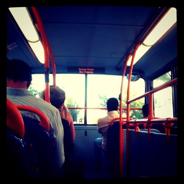 The second floor of a London double-decker bus, with a few travellers looking out of the front window. It's bright and sunny outside.