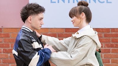 A boy and girl stand outdoors smiling at each other as she touches his shoulder and he holds her hand