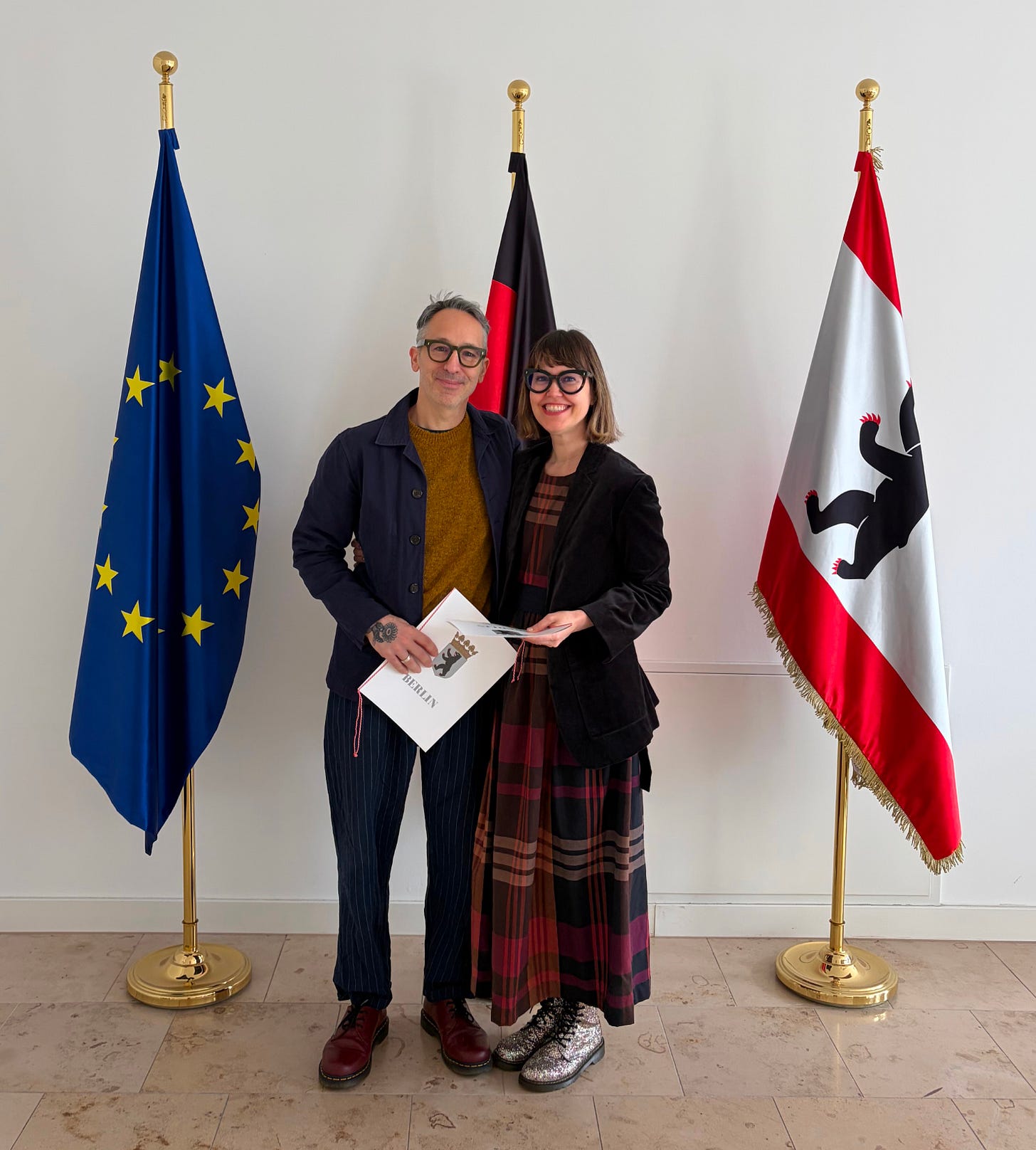 A middle-aged white couple stand in front of the EU, Berlin, and German flags, smiling and holding certificates.