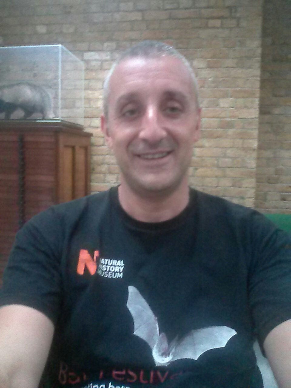 A man with short greying hair and kind eyes smiles at the camera, wearing National History Museum black t-shirt with a bat on it. A man with short greying hair and kind eyes smiles at the camera, wearing National History Museum black t-shirt with a bat on it.